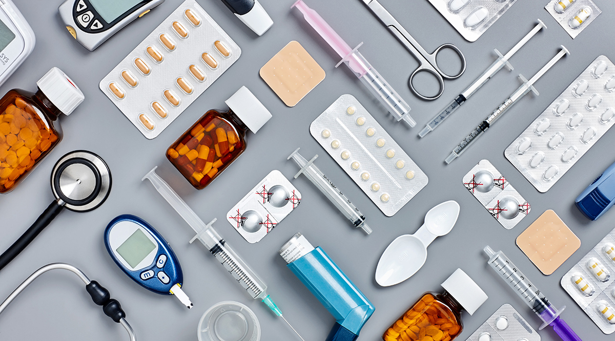 A variety of essential medical supplies including pills, syringes, inhalers, glucose monitors, and first aid tools neatly arranged on a gray surface.